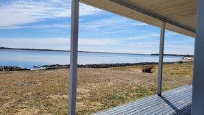 A view from a covered porch overlooking a calm lake with a rocky shoreline and a distant tree line. - Oceanfront cottage with private beach access near Shediac, New Brunswick (Grande-Digue)