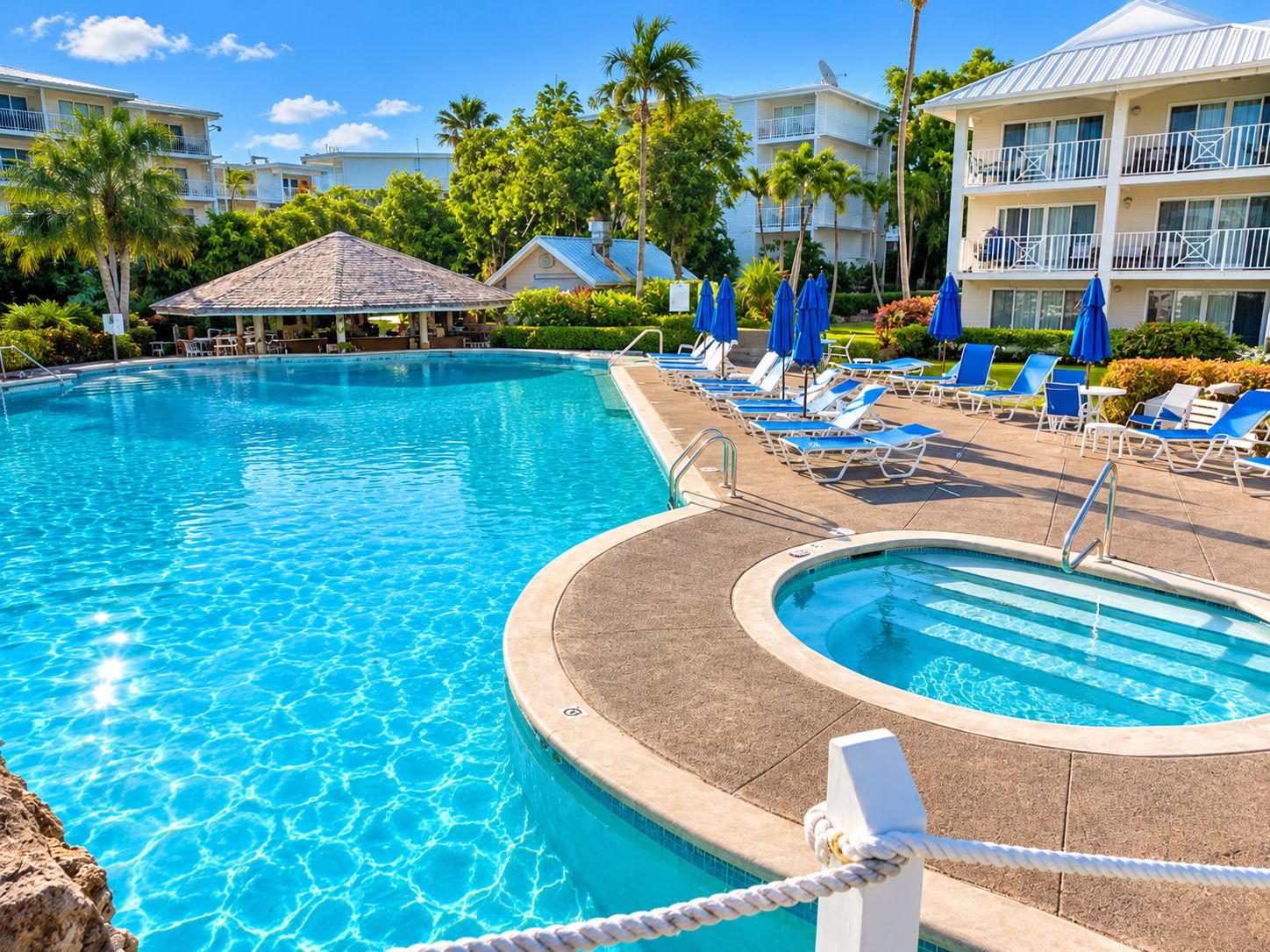 A swimming pool with lounge chairs and umbrellas, surrounded by palm trees and hotel buildings.
