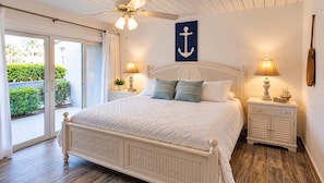 A bedroom with a bed, two bedside tables, a ceiling fan, a view through a glass door, and a wall-mounted anchor decoration.