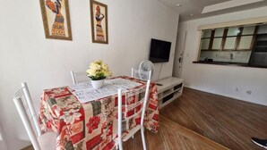 A dining area with a table covered in a floral tablecloth, white chairs, and a vase of flowers. There are two framed artworks on the wall and a flat-screen TV mounted above the table. - SUH Copacabana 105 Metro Posto 5 (Rio de Janeiro)