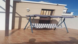 A patio area with a striped outdoor table and chairs, a tiled floor, and a building with a balcony in the background. - Luxury Villa - Sleeps 4, Hottub Jacuzzi & Parking (Tías)