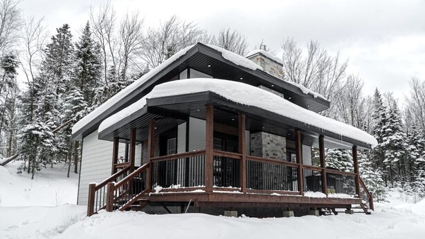 A wooden cabin with a covered porch, surrounded by snow-covered trees.