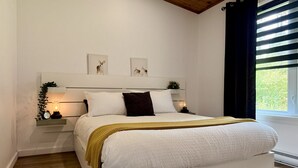 A bedroom with a bed, bedside tables, a nightstand, a window with curtains, and wall-mounted artwork.
