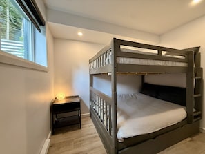 A bunk bed with a wooden frame, a bedside table with a lamp, and a window with blinds. - Zénitude En Forêt Chalet – Spa & Precious Moments (Saint-Damien-de-Buckland)