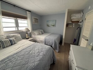 A bedroom with two beds, a nightstand with a lamp, a painting on the wall, and a storage area with folded towels.
