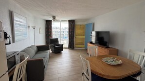 Una sala de estar con sofá, una silla, una mesa de comedor con sillas, televisor y vista a edificios a través de una puerta.