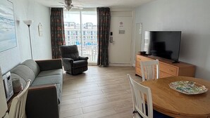 Una sala de estar con un sofá, un televisor, una mesa de comedor con sillas y vistas al exterior.