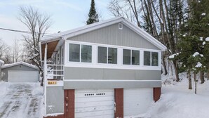 A two-story building with a garage, surrounded by snow-covered ground and trees. - The Grove 1/2 mile from Boyne Mountain Sleeps 4 (Boyne Falls)