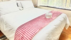 Een net opgemaakt bed met witte linnens en een roze gebreid plaid, een witte handdoek op het bed, en een raam met uitzicht op groen.