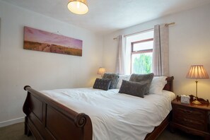 A bedroom with a bed, two bedside lamps, a nightstand, a window with curtains, and a painting on the wall.