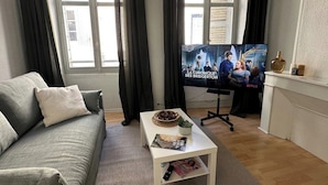 A living room with a sofa, a coffee table with magazines, a TV displaying a show, and a window with curtains. - Serenity - In the heart of the Dax thermal baths (Dax)