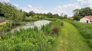 Un sentier paisible le long de la rivière, avec un pont en bois, une végétation luxuriante et un petit bâtiment surmonté d’un toit rouge.