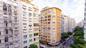 Un edificio residencial de varios pisos con balcones, unidades de aire acondicionado y una calle frente a él con peatones y vehículos.