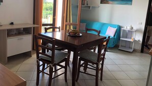 A dining area with a wooden table and chairs, a blue sofa, a painting of a beach, and a small white shelf with a vase.