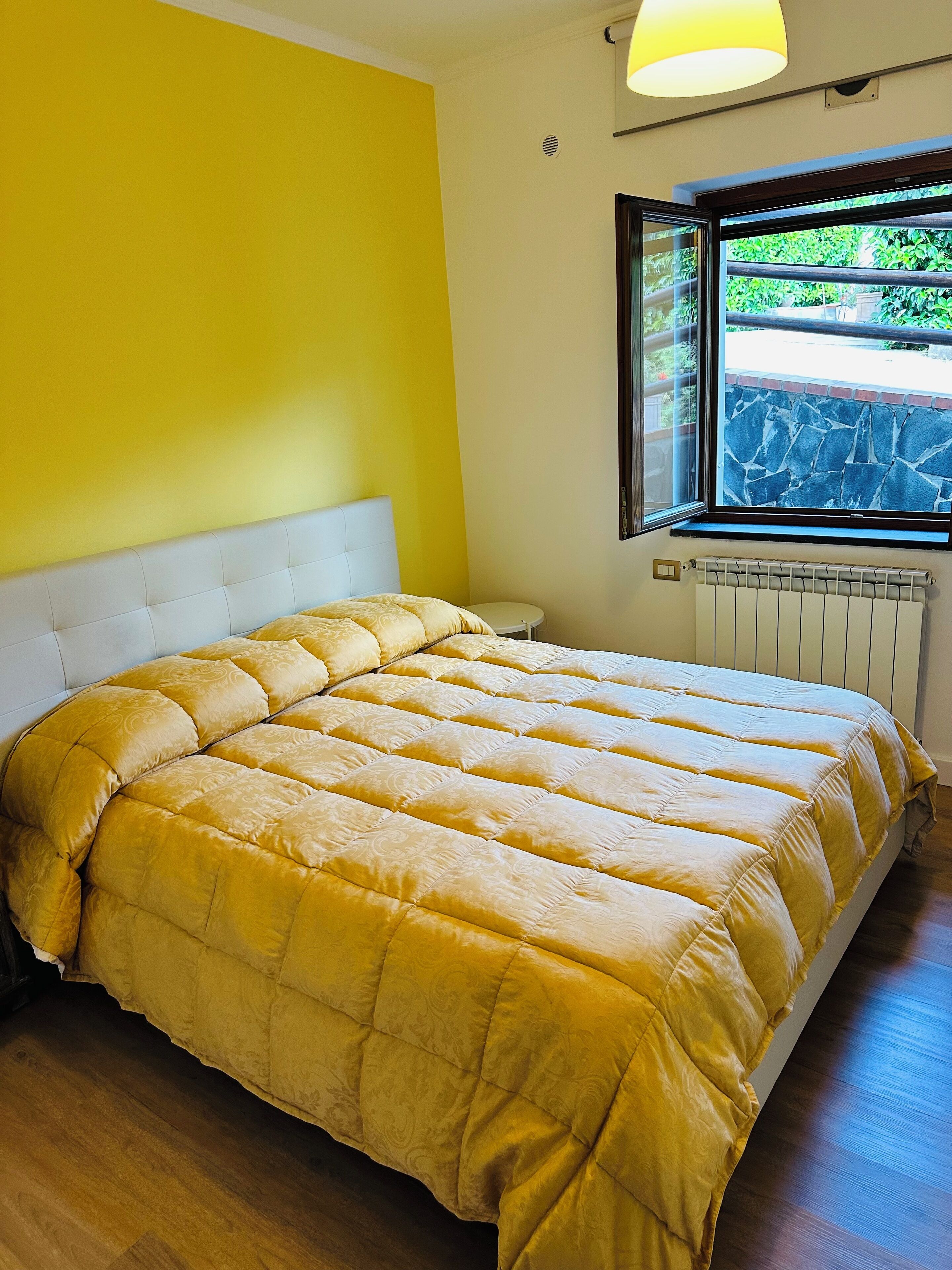 A bedroom with a bed, a window with a view of greenery, and a radiator.