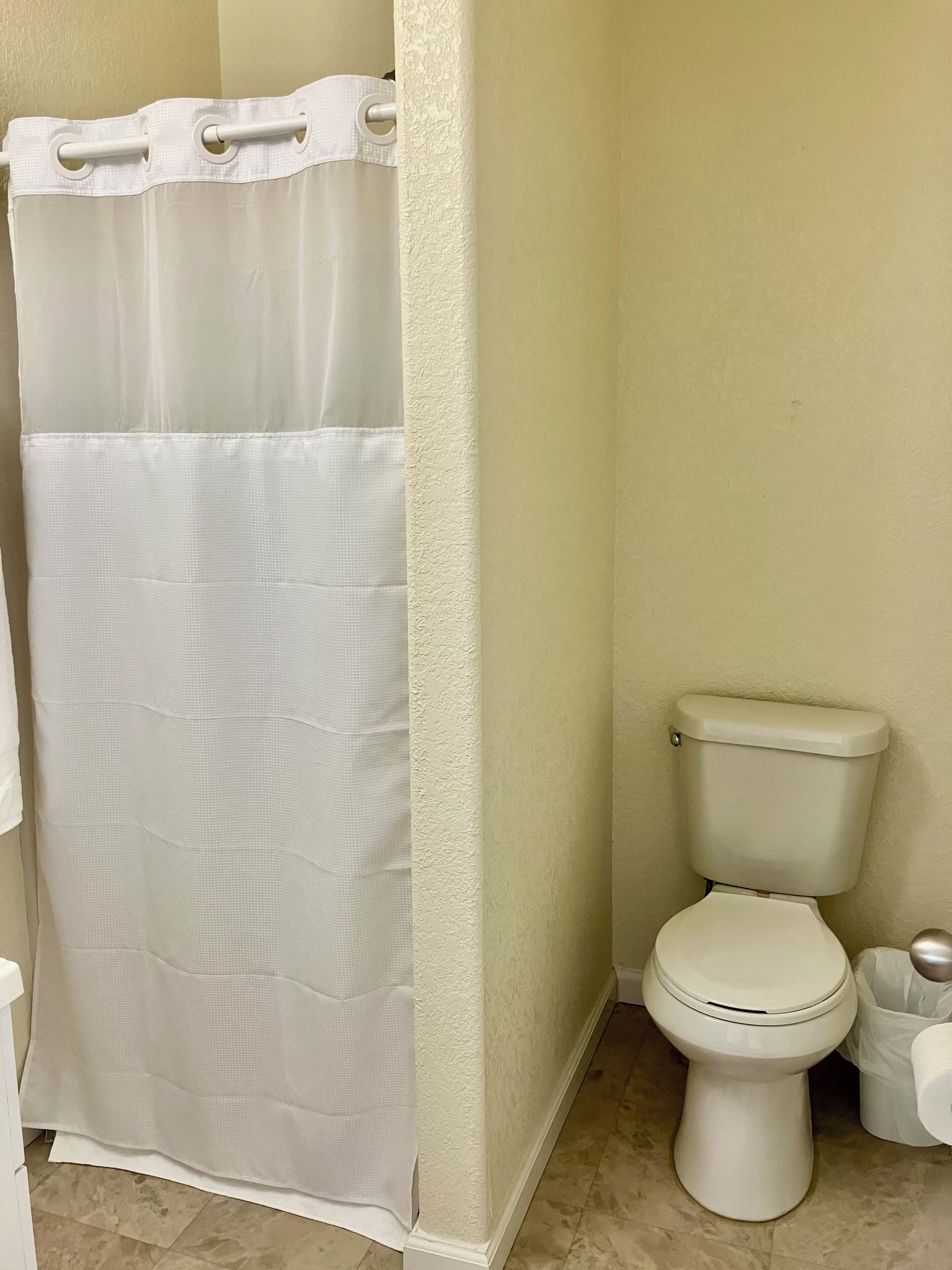 A bathroom with a white shower curtain, a toilet, and a trash bin.