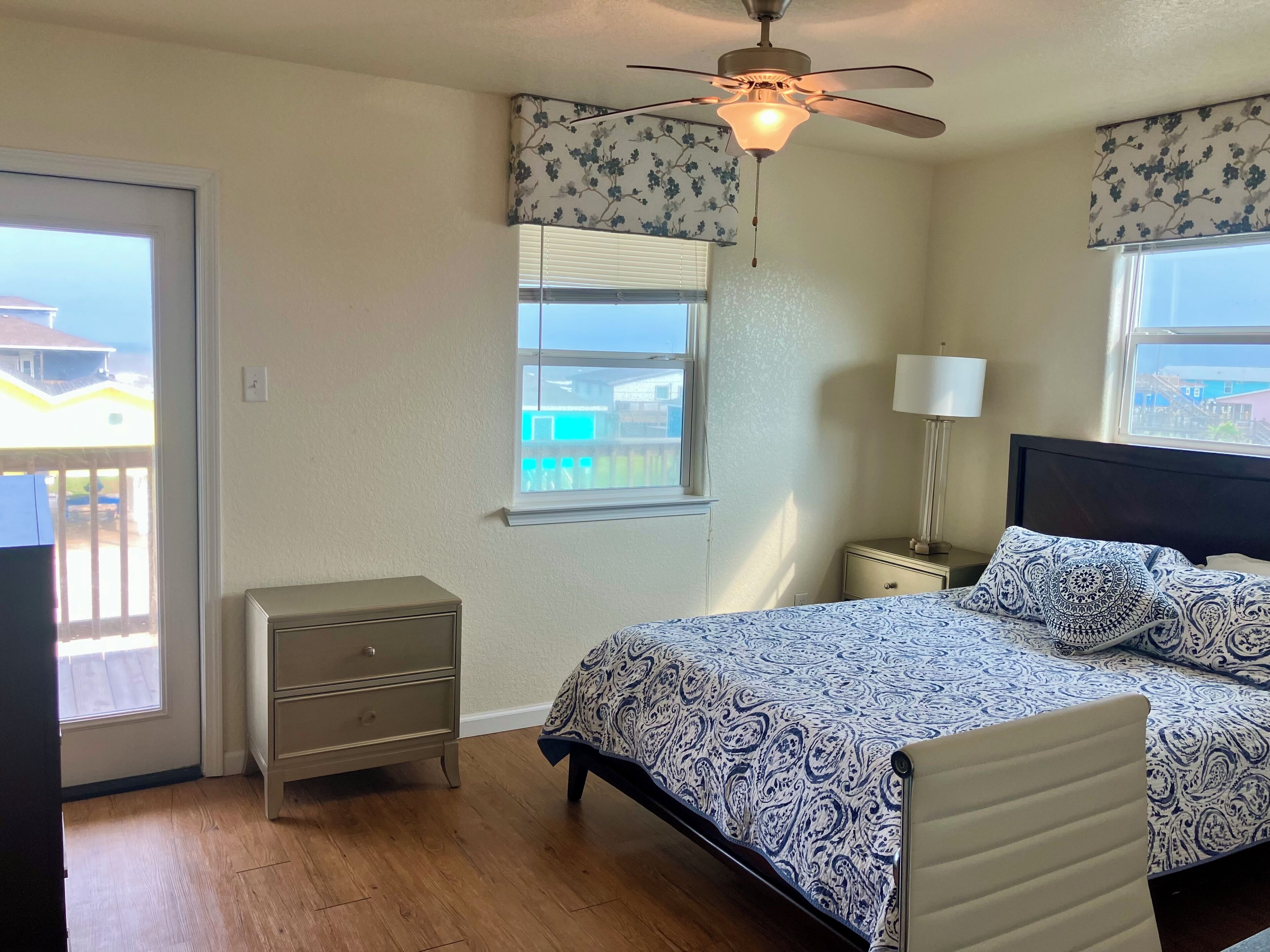A bedroom with a bed, a nightstand, a lamp, a ceiling fan, and a window with a view of a balcony.