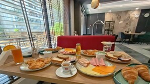 Una tavola imbandita per la colazione con croissant, formaggio, prosciutto e succo d'arancia in una moderna stanza d'albergo con ampie finestre e un divano rosso.