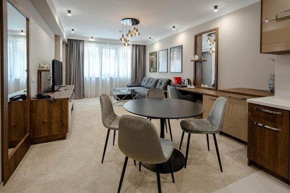 Een moderne hotelkamer met een ronde eettafel, grijze gestoffeerde stoelen, een bank, een televisie en een grote spiegel.
