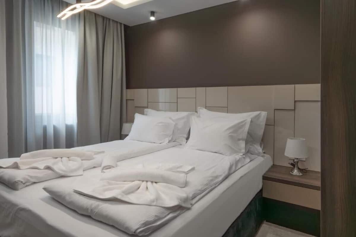 Ein ordentlich gemachtes Bett mit weißen Leinentüchern und Kissen, ein Nachttisch mit Lampe sowie ein Fenster mit durchsichtigen Vorhängen.