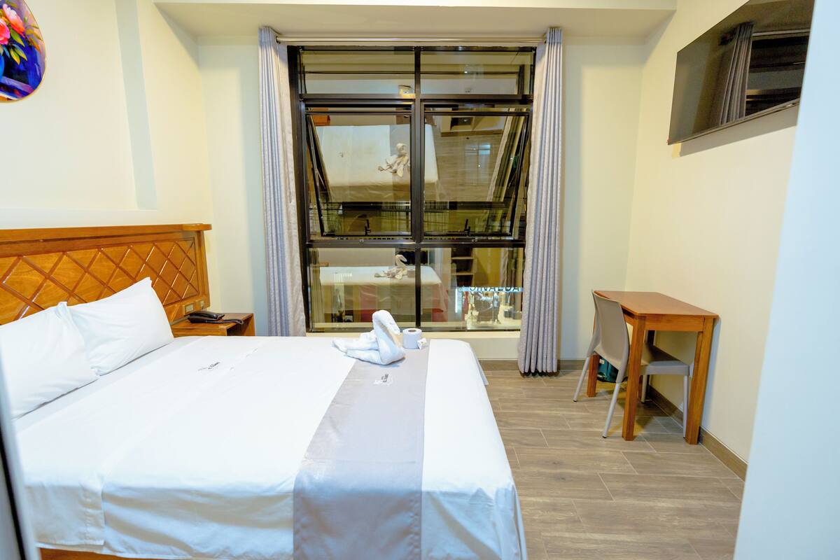 A hotel room with a bed, a wooden headboard, a small table, and a window with curtains.