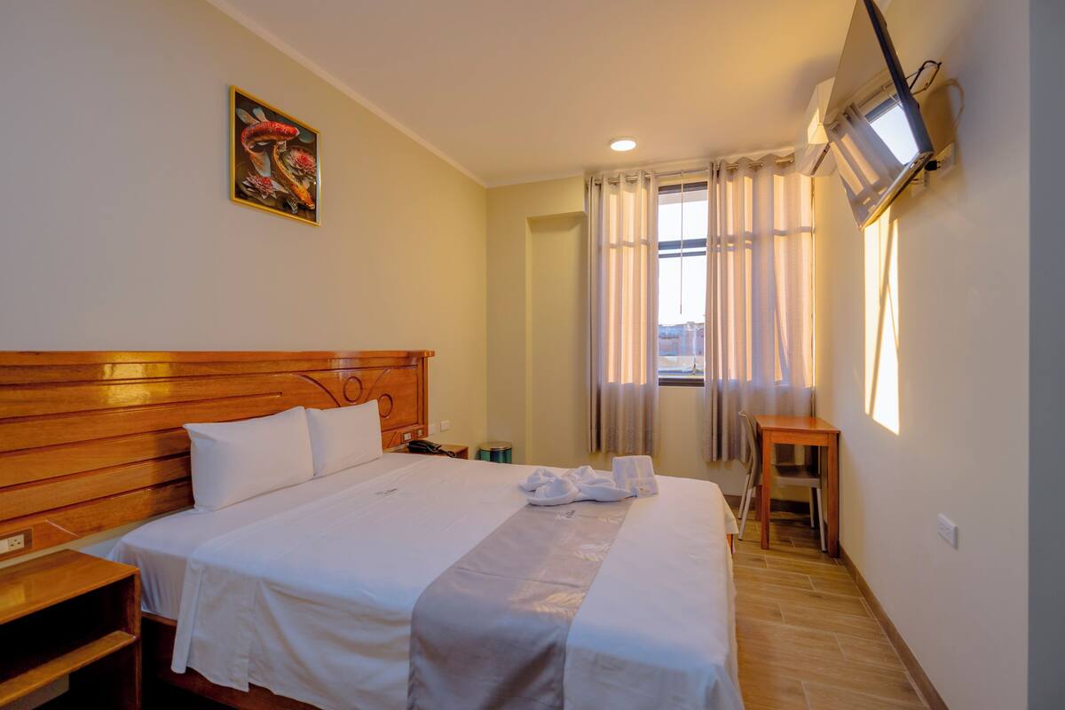 A hotel room with a wooden headboard, a bed with white linens, a small table, a TV mounted on the wall, and a window with curtains.