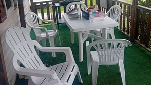 A covered patio with white plastic chairs and a table, surrounded by a wooden deck and a view of a grassy area.