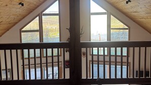 A high-ceilinged attic room with wooden beams, a balcony with a white railing, and large windows offering a view of the outdoors.