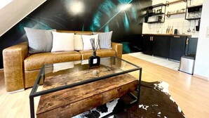 Una sala de estar moderna con una mesa de centro de vidrio, un sofá de cuero marrón y un tapete de piel de vaca. Se ve una cocina al fondo con estanterías de madera y encimera negra.