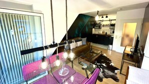 Un espacio habitable moderno con una mesa de comedor de cristal, sillas moradas, una mesa de centro de cristal, un tapiz de cuero de vaca, un sofá de cuero y una zona de cocina con un tapiz de cuero de vaca blanco y negro.