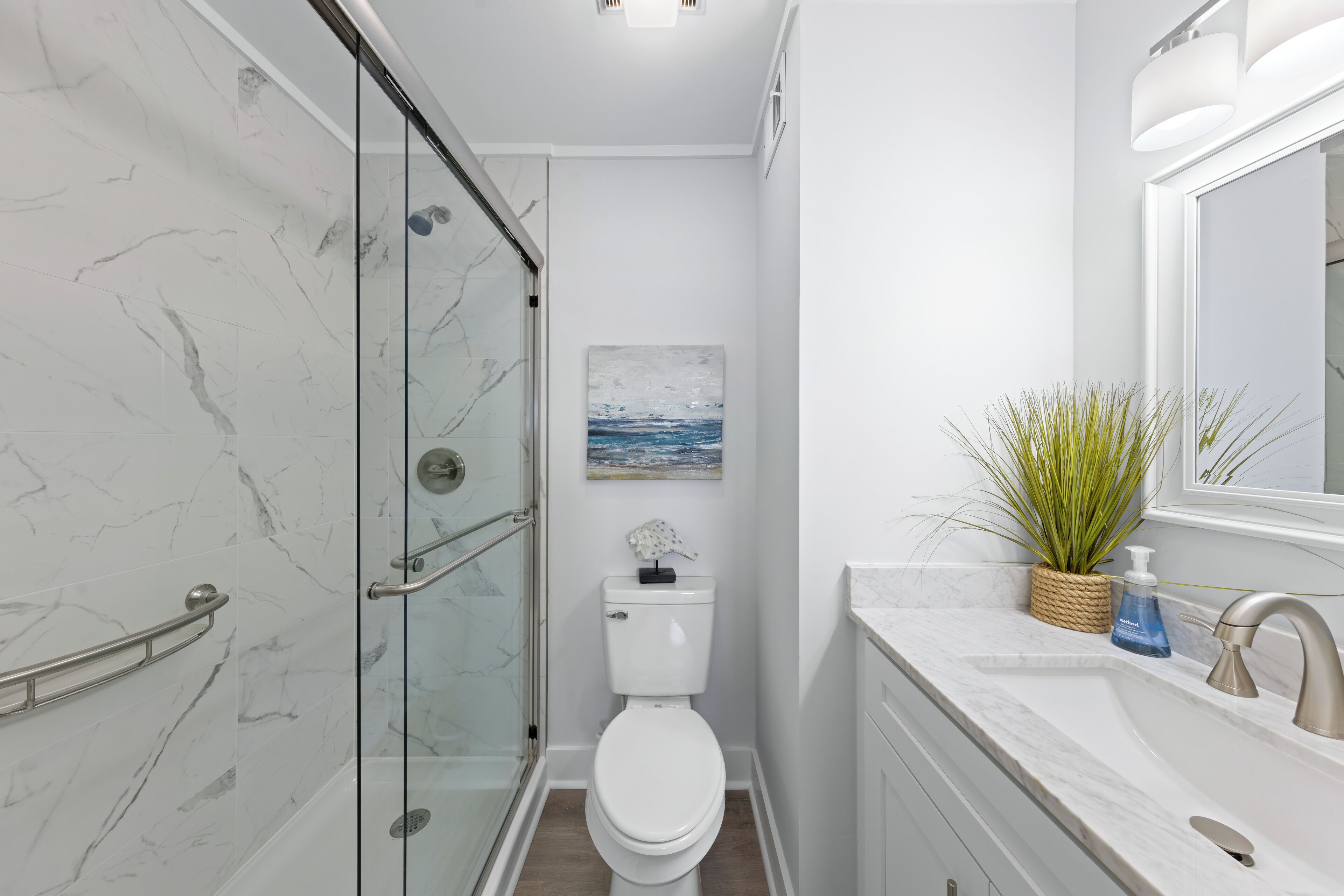 A bathroom with a glass shower enclosure, a toilet, a sink with a marble countertop, and a potted plant.