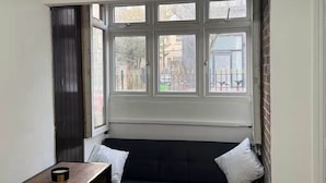 A corner room with a dark sofa, a wooden table, and a view through large windows.