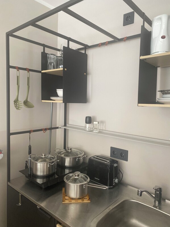 Una cocina compacta con un lavamanos, dos ollas de acero inoxidable, un tostador y utensilios de cocina colgantes.