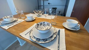 A dining table set with white and patterned tableware, including bowls, plates, and cutlery, with a wooden table and a kitchen in the background.