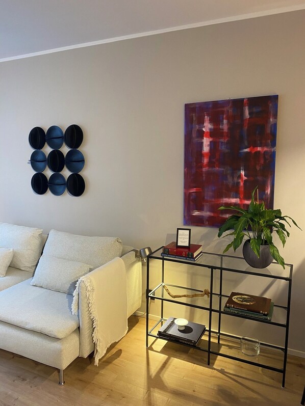 Una sala de estar con un sofá blanco, una mesita auxiliar de metal negro con libros y una planta, un objeto circular montado en la pared y un cuadro abstracto de gran tamaño.