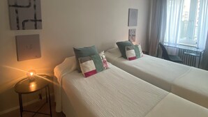 A hotel room with two single beds, a small round table, a desk, and a window with curtains.
