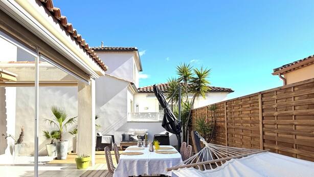 Une terrasse en bois avec un hamac, une table recouverte d’une nappe blanche et des plantes en pot.