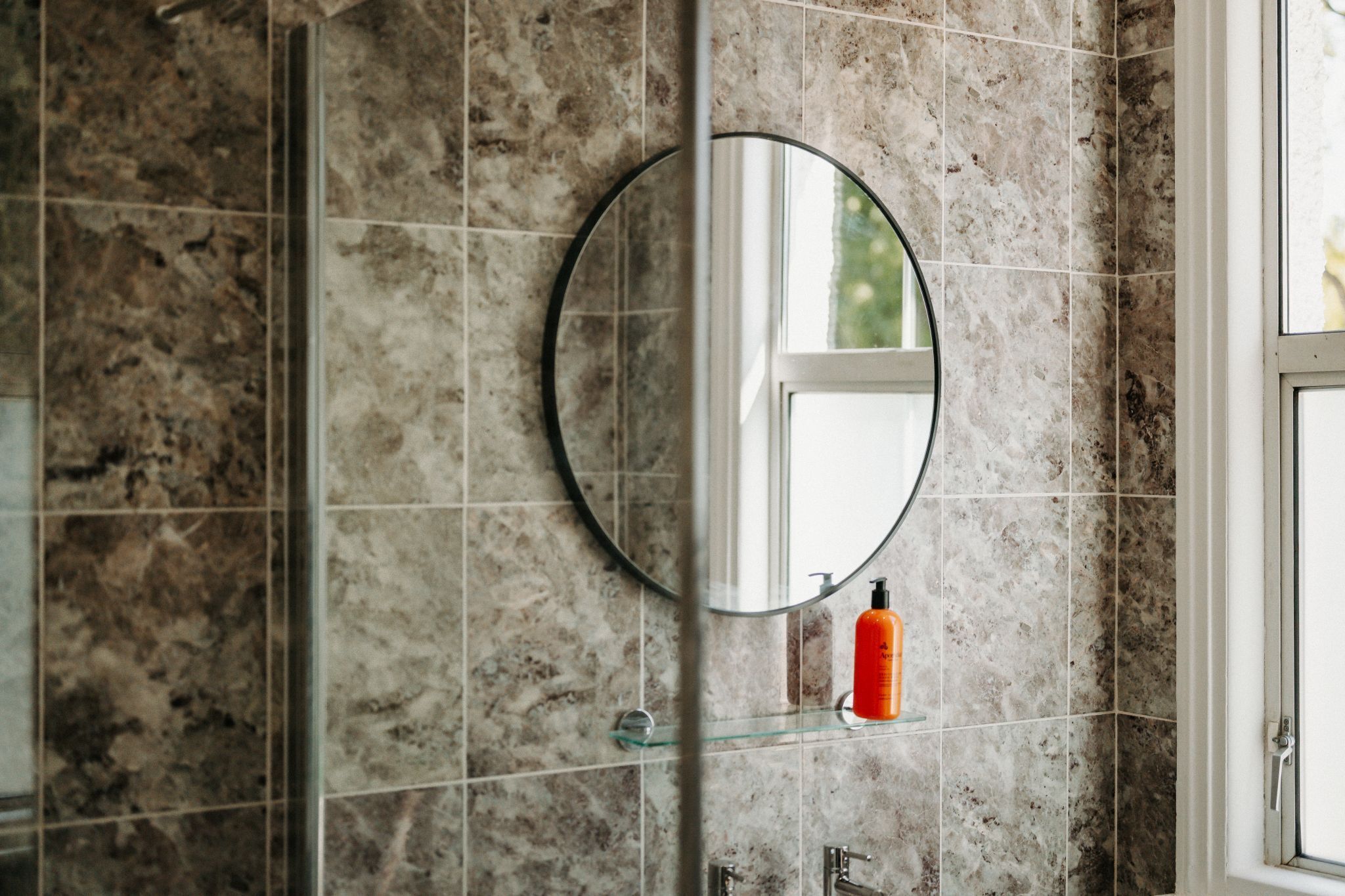 A bathroom with a round mirror, a glass shelf, and a window.