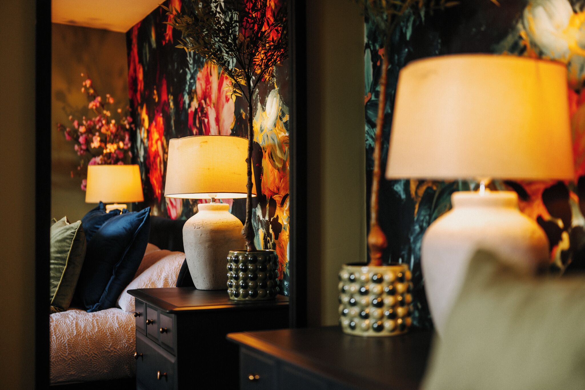 A bedroom with a bed, two bedside tables with lamps, a decorative vase, and a floral wall mural.