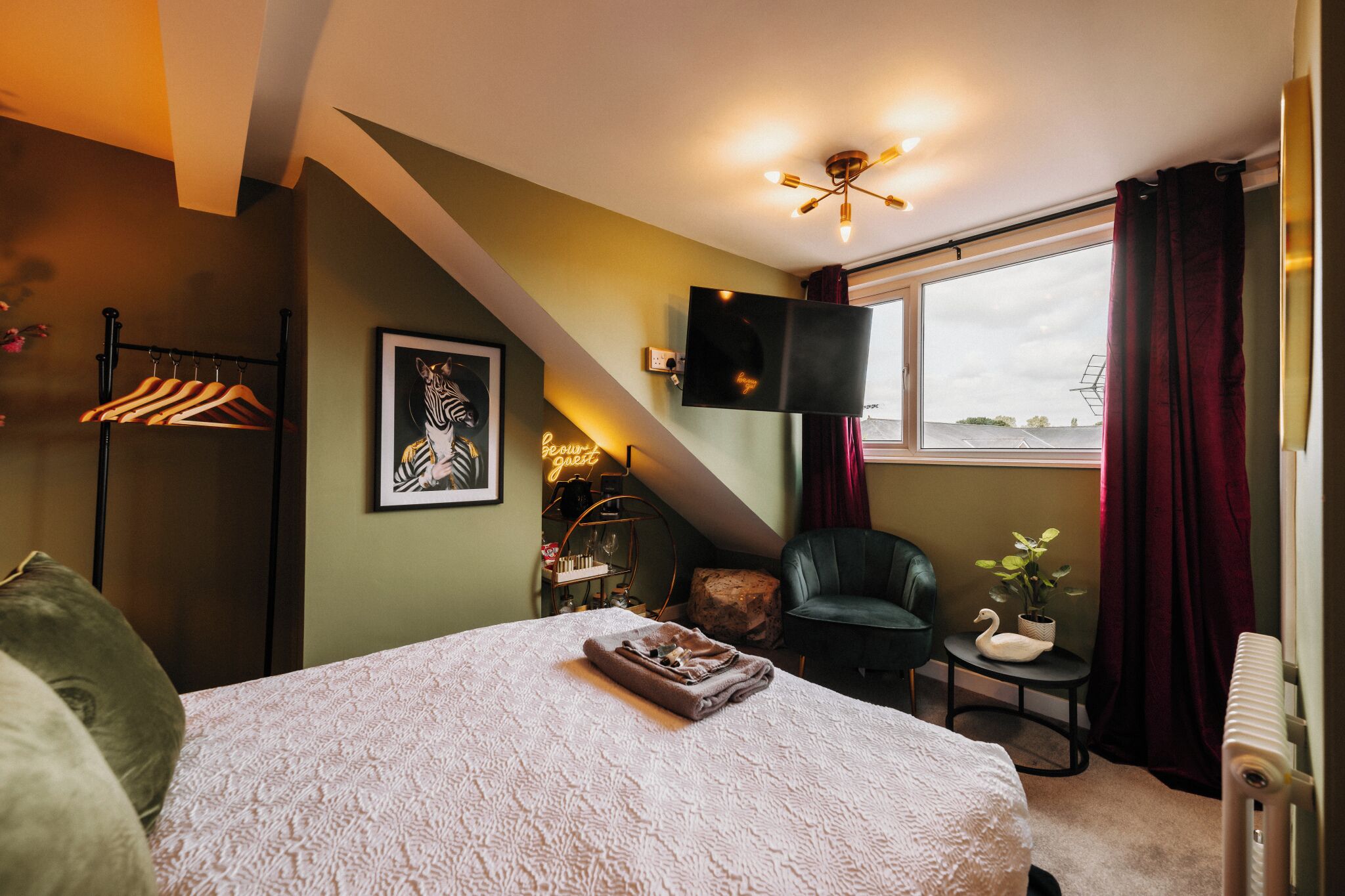 A bedroom with a bed, a TV mounted on the wall, a window with curtains, a chair, and a small table with a plant.