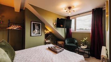 A bedroom with a bed, a TV mounted on the wall, a window with curtains, a chair, and a small table with a plant.
