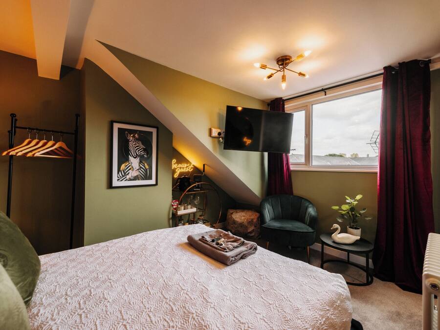 A bedroom with a bed, a TV mounted on the wall, a window with curtains, a chair, and a small table with a plant.