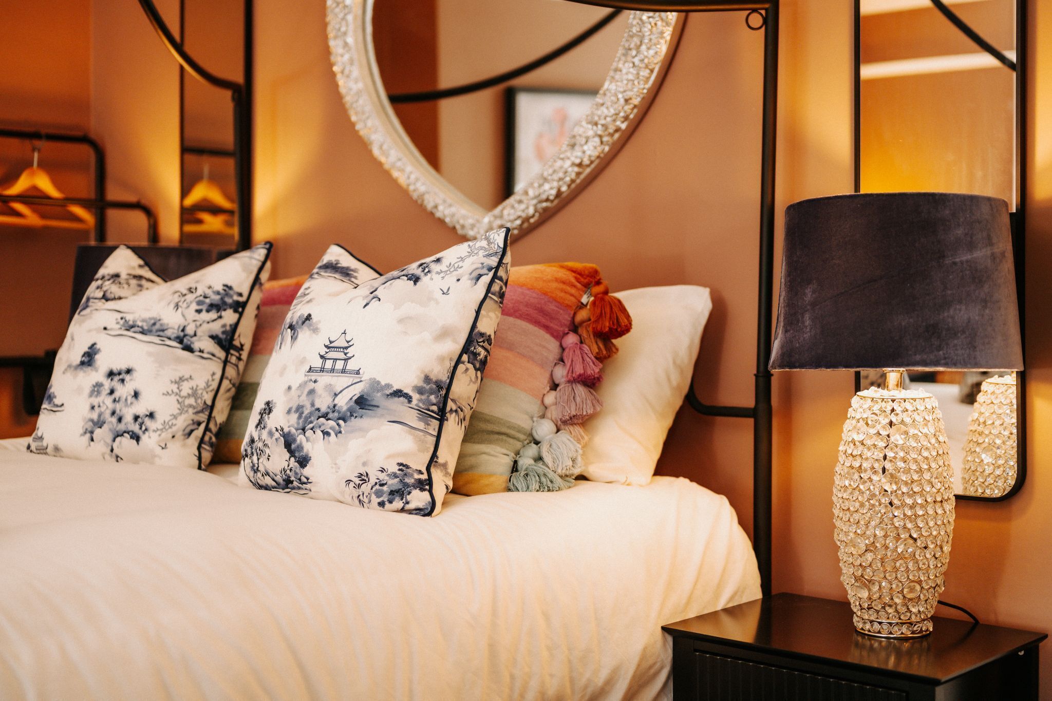 Un lit bien fait, agrémenté d’oreillers à motifs floraux, d’une lampe décorative et d’un miroir posé sur une table de chevet.