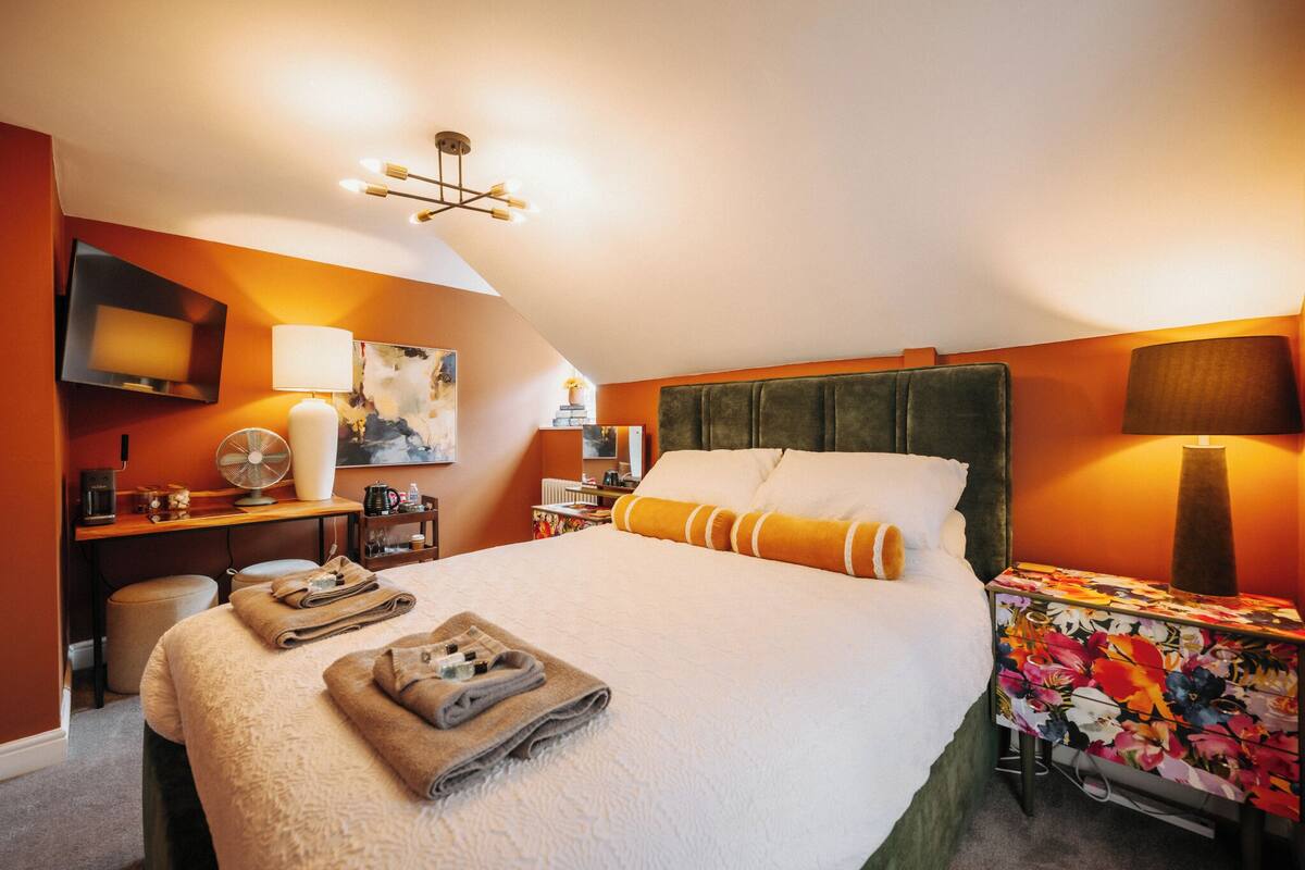Ein Hotelzimmer mit einem großen Bett, einem Flachbildfernseher, einem Schreibtisch mit Lampe, einem Ventilator und einer Tischdecke mit Blumenmuster.