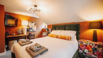 A hotel room with a large bed, a flat-screen TV, a desk with a lamp, a fan, and a floral tablecloth.