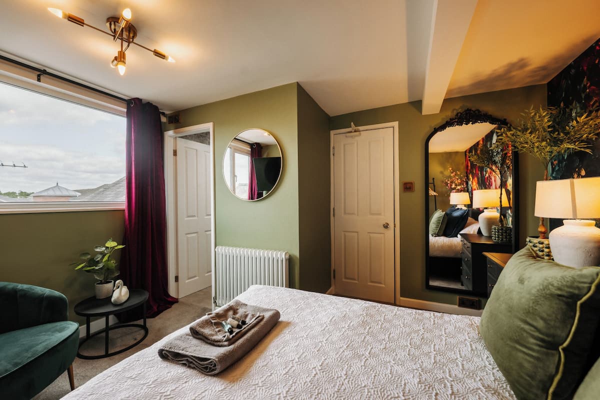 Ein Schlafzimmer mit einem Bett, einem runden Spiegel, einer Heizung, einem Fenster mit Vorhängen, einem grünen Sessel und einer Topfpflanze.