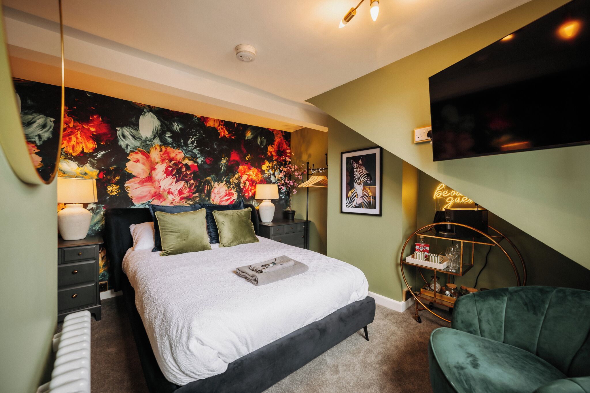 A bedroom with a large floral mural, a bed with a white comforter, a green armchair, and a neon sign.