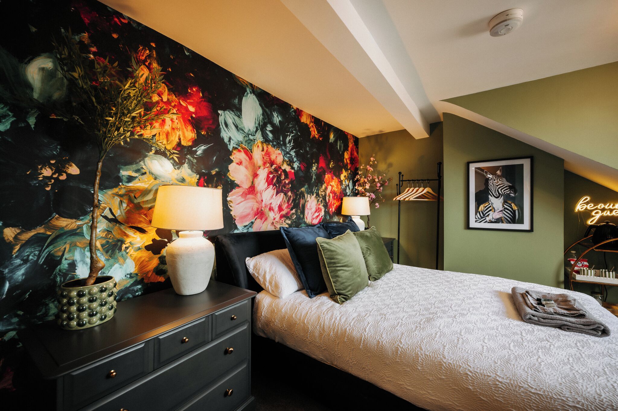 A bedroom with a floral mural, a bed with a white comforter, two bedside lamps, a dresser, and a neon sign.