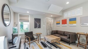 Un salon moderne avec un canapé en cuir marron, une table basse en bois, une chaise noire et une petite table ronde. Un grand miroir orne le mur, il y a une fenêtre avec des stores et des œuvres d’art sont accrochées au mur.