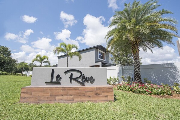 Une enseigne portant l’inscription « Le Rêve » devant un bâtiment moderne, avec une pelouse verdoyante et des palmiers.